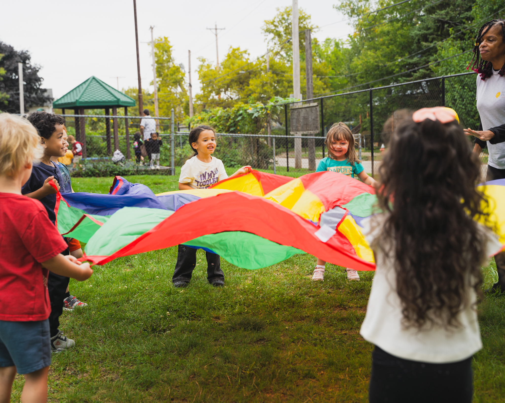 Children and teachers playing with a colorful parachute on the grassy field at Cleveland Child Care