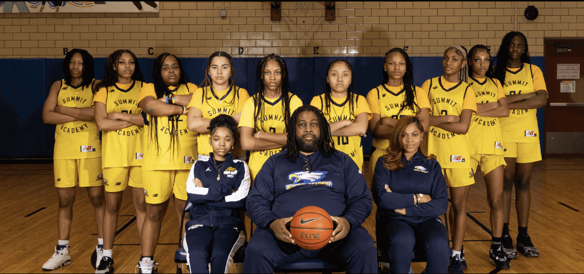 A basketball team poses in their yellow uniforms with coaches in front holding a basketball.
