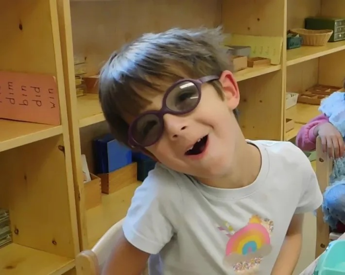 A smiling child in glasses standing in front of the classroom shelves