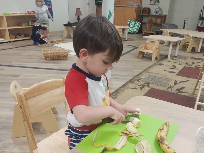 A child at Montessori Children's Academy slicing a banana on a green cutting board during practical life work