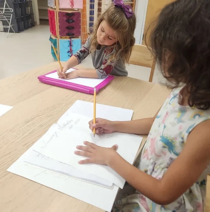 Two children practising handwriting together with pencils and paper at a wooden desk